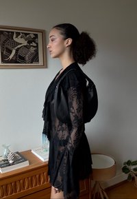 Robe en dentelle noire avec détails à volants, manches longues et corsage ajusté. Complétée par un sac à main en cuir noir. Textures douces et design transparent.