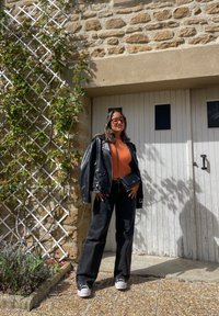 Zwarte leren jas, oranje gestructureerde trui, zwarte jeans, grijze sneakers. Achtergrond bevat een stenen muur en witte houten deuren met zwarte ramen.