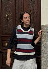Navy, grey, and burgundy striped knit vest with a ribbed hem. Styled with a black handbag and a layered black long-sleeve top.
