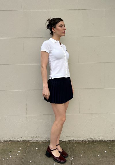 Camisa polo blanca de manga corta con botones al frente, combinada con una falda plisada azul marino y zapatos de tacón cuadrado color burdeos.