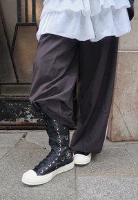 Personne portant des bottes montantes Converse noires avec des anneaux métalliques, un pantalon noir ample et une chemise blanche superposée, debout sur un pavé en pierre.