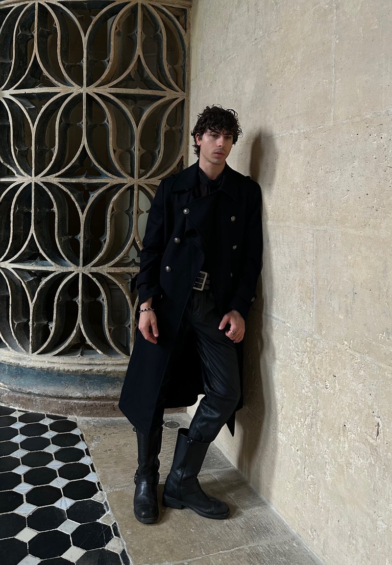 Jeune homme aux cheveux bouclés portant un manteau noir, un pantalon en cuir et des bottes, appuyé contre un mur en pierre beige avec des carreaux décorés à proximité.