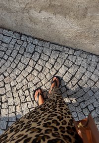 Sandale negre pe picioare goale, asortate cu o rochie cu imprimeu de leopard până la genunchi, stând pe un paviment de piatră cubică texturată.