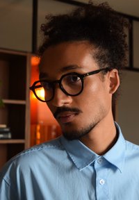 Jeune homme aux cheveux bouclés relevés, portant des lunettes rondes noires et une chemise à boutons bleu clair, à l'intérieur avec un éclairage chaleureux.