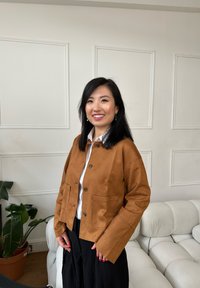 Veste courte en daim marron à la silhouette structurée, avec de grandes poches, une fermeture à boutons et des manches larges, portée avec une chemise blanche.