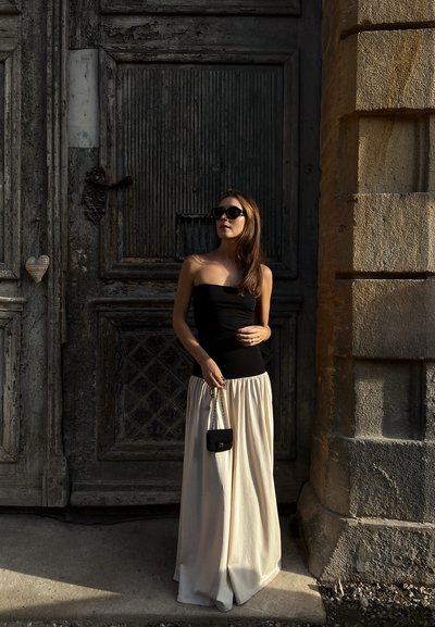 Top negro sin mangas combinado con una falda larga y fluida de color crema. Un pequeño bolso negro complementa el conjunto, situado frente a una puerta de madera texturizada.