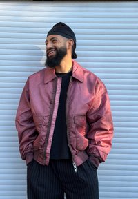 Rdeča satenasta bomber jakna z ovratnikom, dvema sprednjima žepoma in rebrastimi manšetami, nošena čez črno srajco in črtaste hlače.