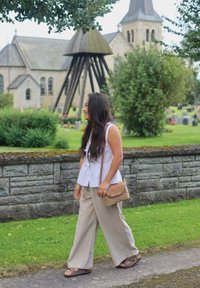 Heledhalli kivi sein, mille ees on roheline muru, ja sellele on toetatud valges peplum-särgis ja beežides laia säärega pükstes inimene, kes kannab väikest kootud kotti.