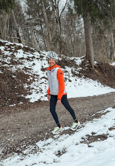 Mujer con ropa de invierno estirando la pierna en un sendero nevado rodeado de árboles sin hojas y bosque.