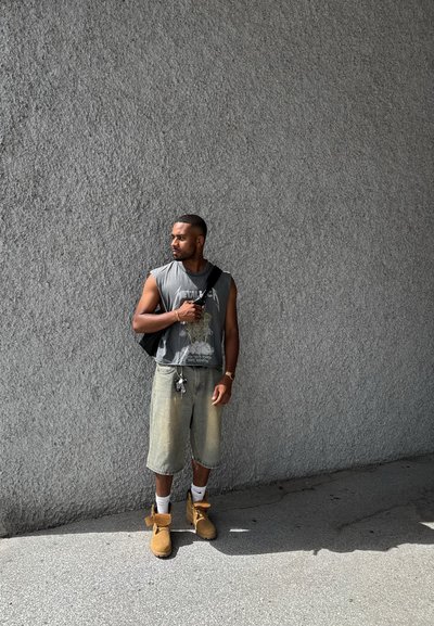 Camiseta gráfica sin mangas de color gris, pantalones cortos de cargo gris claro, botas amarillas y calcetines blancos. Mochila negra y llavero visibles. Fondo de pared texturizada gris.