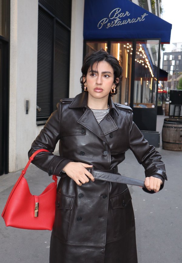 Femme ajustant la ceinture de son manteau en cuir noir, portant un sac à main rouge, debout sur un trottoir en ville près d'un bar-restaurant avec une toile bleue et des guirlandes lumineuses.