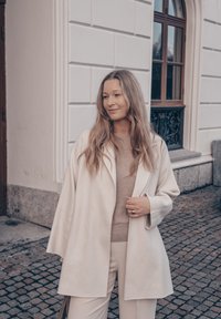 Manteau beige clair au design décontracté, en tissu de laine, manches larges et revers crantés, porté sur un pull beige doux et un pantalon crème.