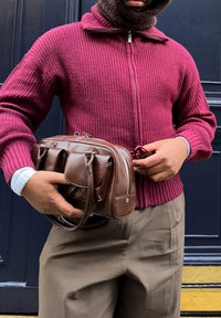 Man wearing a maroon knitted jumper and brown trousers holding a brown leather crossbody bag and adjusting glasses in front of a dark door.