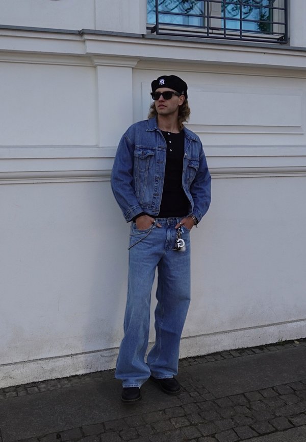 Giacca di jeans e jeans azzurri, maglia nera a maniche lunghe, berretto nero e occhiali da sole. Presenta dettaglio a catena sui jeans.