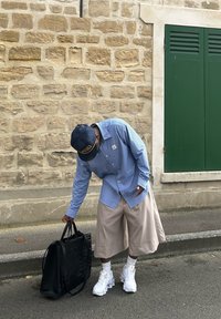 Ρυθμισμένο πουκάμισο με μπλε και λευκές ρίγες, μπεζ σορτς, λευκά chunky sneakers και μια μαύρη τσάντα tote, με υφασμάτινα σχέδια στα ρούχα.