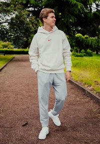 Λευκό φούτερ Nike με κόκκινο λογότυπο, γκρίζα παντελόνια φόρμας και λευκά αθλητικά παπούτσια. Το μοντέλο στέκεται σε ένα χωμάτινο μονοπάτι περιτριγυρισμένο από πράσινα φυτά.