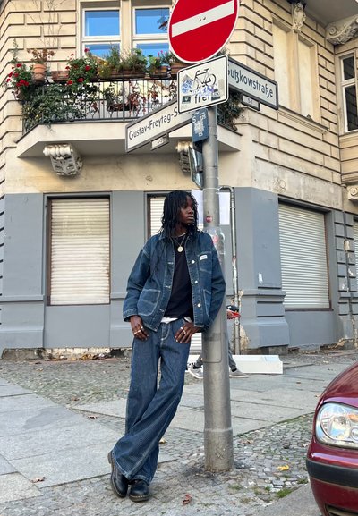 Conjunto de chaqueta y pantalones de mezclilla en azul oscuro con rayas verticales. Camisa negra debajo. Botas negras. Calle urbana con señalización y detalles de edificios.