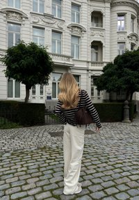 Prugasti crno-bijeli pulover, krem hlače širokih nogavica, sivi tenisice i tamnosmeđa torba naspram pozadine ukrašene bijele zgrade.