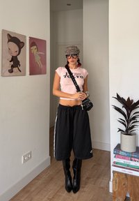 Pantaloni scurți atletici negri cu dungi albe, tricou roz scurt, cizme negre până la genunchi și o șapcă tip șoferailă în carouri. Geantă mică neagră.