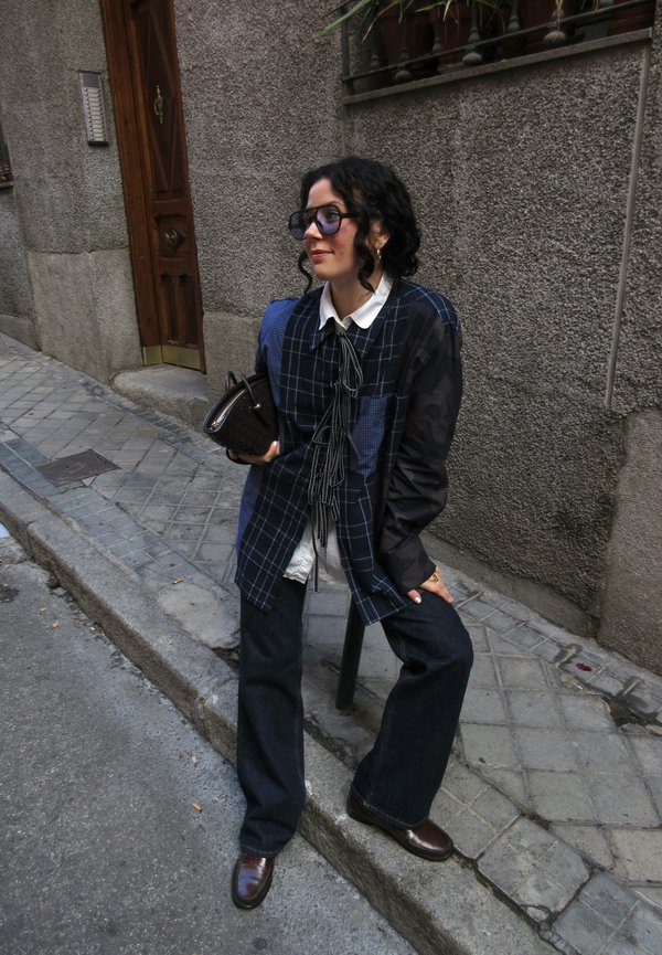 Camicia quadrata blu oversize con maniche nere, colletto bianco e dettaglio della cravatta nera. Pantaloni svasati in denim scuro e scarpe marroni. Borsa marrone.
