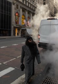 Personne en manteau gris long, chapeau noir et lunettes de soleil marchant près d'une grille de rue fumante à côté d'une camionnette blanche et d'un bâtiment historique avec des drapeaux américains.