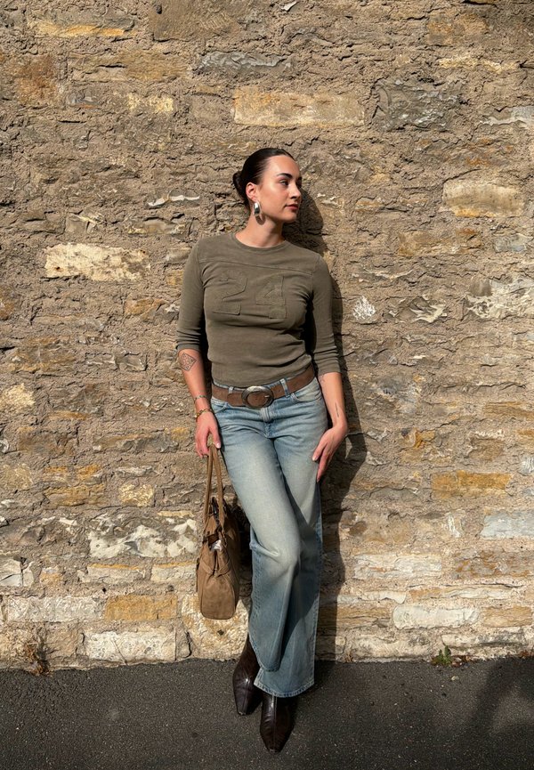 Mujer con el cabello recogido en un moño que se apoya contra una pared de piedra texturizada, lleva una camiseta verde oliva, vaqueros azul claro, botas oscuras, pendientes plateados y sostiene un bolso marrón claro.