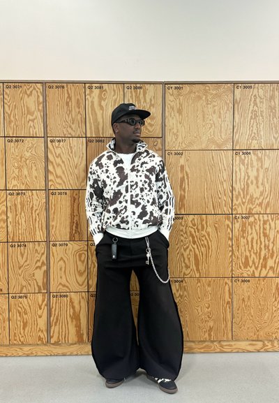 Chaqueta con estampado de vaca en blanco y negro, con cremallera y cuello alto, pantalones negros anchos, cadena adjunta y una gorra negra con texto. Taquillas de madera al fondo.