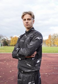 Svart atletisk träningsdräkt med abstrakta grå mönster, dragkedjejacka och elastisk midjeresår, stående på en röd löparbana med gräs i bakgrunden.