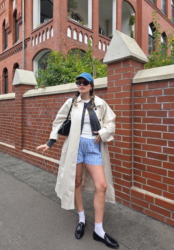 Beige Trenchcoat über einem grauen Cardigan, weißem T-Shirt und blauen gestreiften Shorts. Schwarze Loafers und weiße Socken, mit einer blauen Kappe und Sonnenbrille.