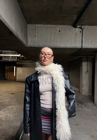 Femme avec des lunettes portant un manteau en cuir noir, une écharpe blanche moelleuse et une robe imprimée, debout dans un parking en béton faiblement éclairé.