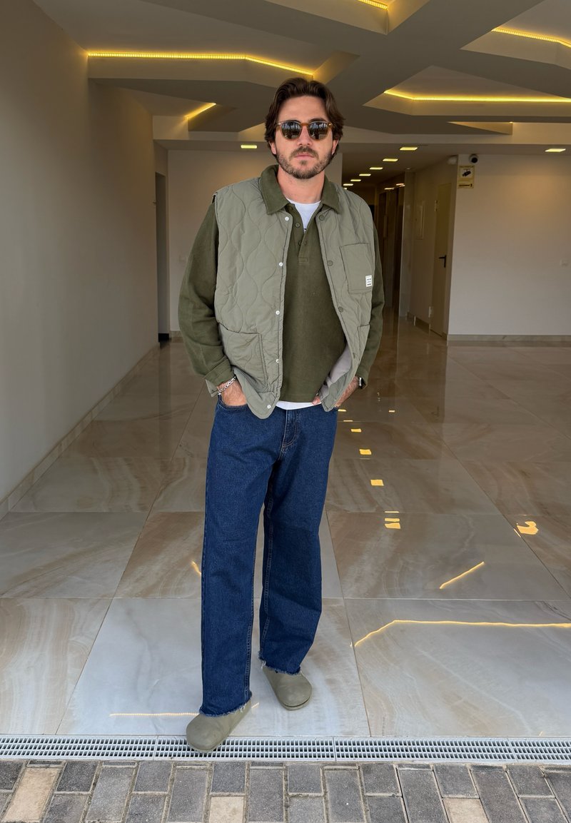 Groene gewatteerde vest over een groen shirt, gecombineerd met wijde blauwe jeans en olijfgroen schoeisel. Gelegen in een moderne interieur.