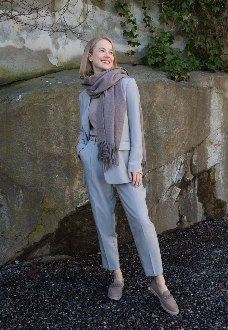 Femme portant un costume gris clair, une écharpe beige et des mocassins beiges, souriante, debout contre un mur de pierre recouvert de lierre.