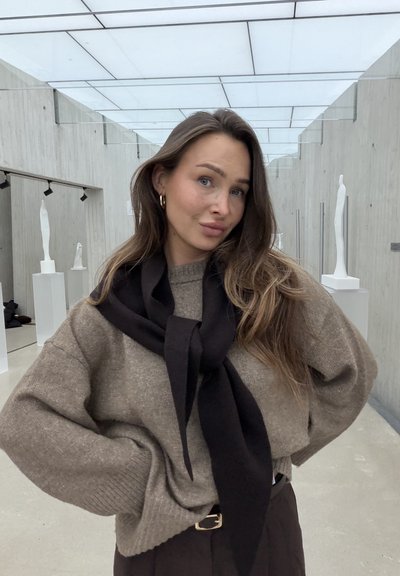 Jersey de punto marrón claro con mangas extragrandes, combinado con una bufanda negra anudada al frente. Pantalones negros de cintura alta. Fondo de una galería moderna.