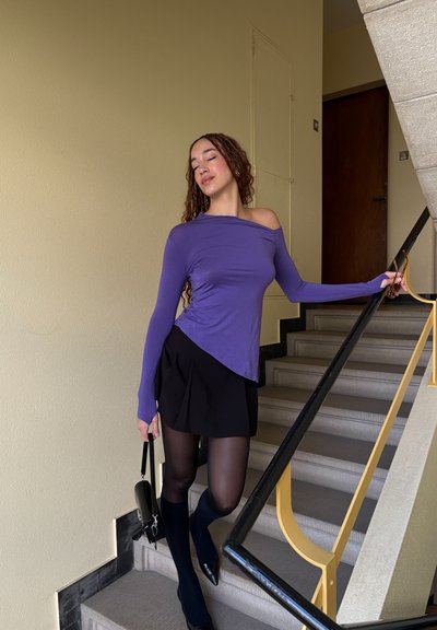 Top de manga larga morado con corte asimétrico, combinado con una falda negra plisada, medias negras transparentes y calcetas hasta la rodilla. Lleva un bolso negro.