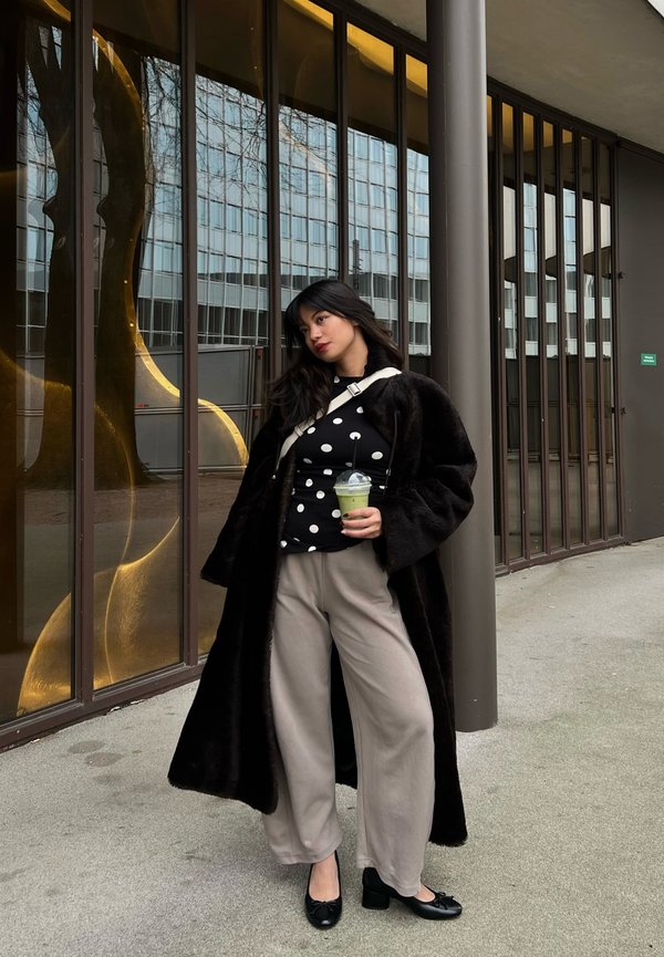 Kobieta w czarnym futrzanym płaszczu i bluzce w groszki trzymająca zielony napój, stojąca obok szklanego budynku z pionowymi brązowymi ramami.