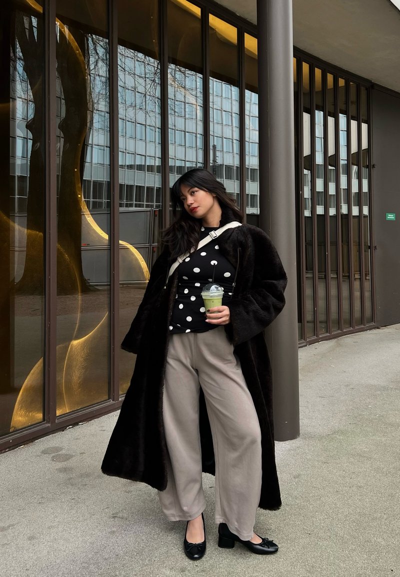 Femme en manteau de fourrure noir et haut à pois tenant une boisson verte, debout près d'un bâtiment en verre avec des cadres verticaux bruns.