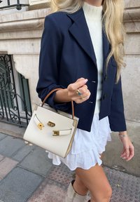 Μικρή κρεμ τσάντα με χρυσές λεπτομέρειες και ανοιχτό καφέ αποχρώσεις. Φοριέται με μπλε σακάκι και λευκή φουστάνα με βολάν, σε φόντο από πέτρα.