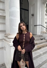 Top tricotat cu nervuri maro, cu accente de butoane aurii, fustă mini neagră și o haină întunecată. Geantă transparentă cu mărgele și curea din lanț. Cadru urban.