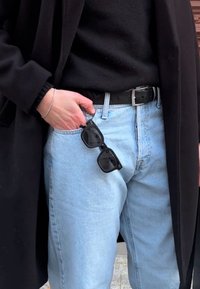 Černé sluneční brýle ležící na světle modré džínové kapse. Osoba má na sobě �černý svetr pod otevíracím černým kabátem a černý pásek.
