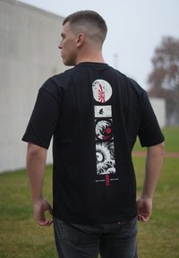T-shirt noir avec un motif graphique en blanc et rouge, représentant des vagues et un motif de soleil au dos, manches courtes et coupe décontractée.