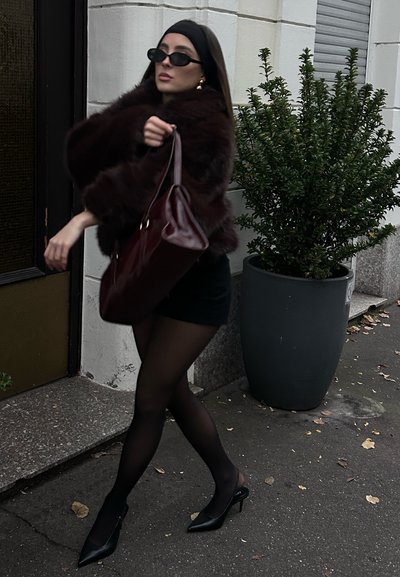 Abrigo de piel marrón, minifalda negra, medias negras con estampado, tacones negros puntiagudos, gafas de sol negras de gran tamaño, sosteniendo un bolso burdeos.