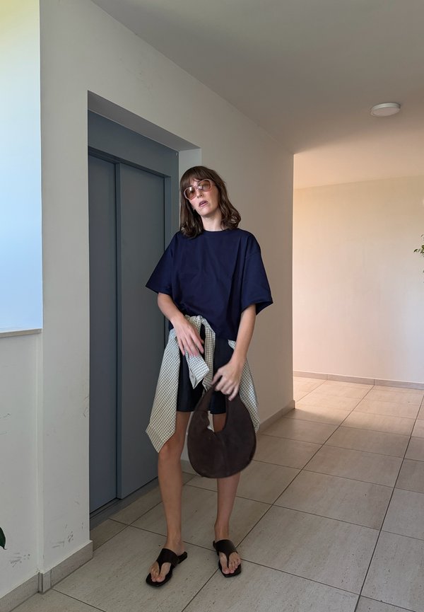 Femme portant des lunettes de soleil, chemise surdimensionnée marine, short noir, tenant un sac marron, debout près d'un ascenseur gris dans un couloir carrelé.