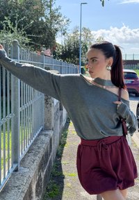 Grå off-the-shoulder tröja med långa ärmar, parat med burgundy shorts med dragsko. Modellen håller en väska och står nära ett staket.