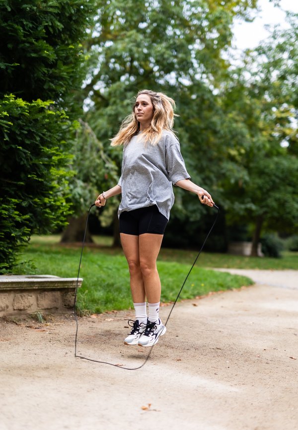 Camiseta oversized gris claro, pantalones cortos de ciclismo negros, calcetines deportivos blancos. Sosteniendo una cuerda para saltar, postura de salto, fondo de parque verde.