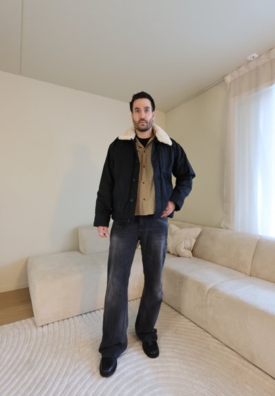 Hombre de pie en la sala de estar con chaqueta negra con cuello de borreguito, camisa beige a cuadros, jeans negros desgastados y zapatos negros.