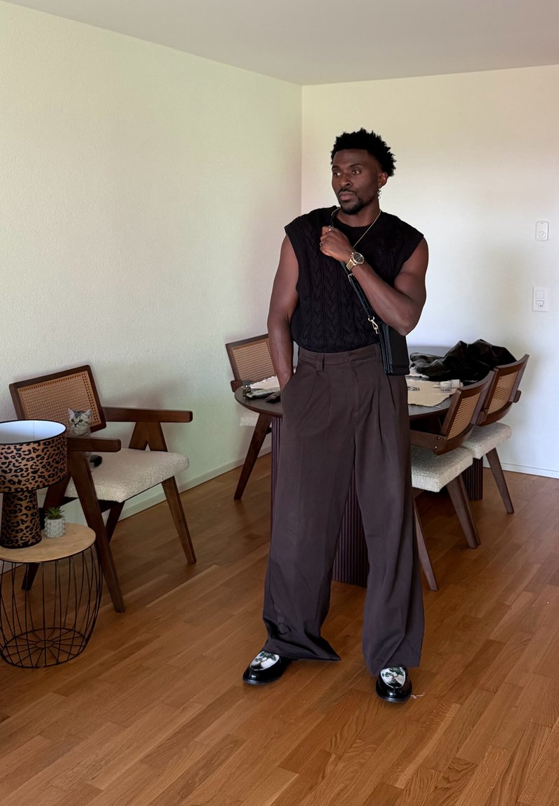 Homme portant un pull sans manches noir et un pantalon large marron se tient dans une salle à manger avec un sol en bois et des chaises, une lampe à motif léopard à proximité.