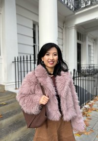 Hellrosa gefälschte Pelz-Bauchtjacke mit weitem Kragen, kombiniert mit einer braunen Ledertasche und einem braunen Wildlederrock. Treppen und ein Eisen-Geländer im Hintergrund.