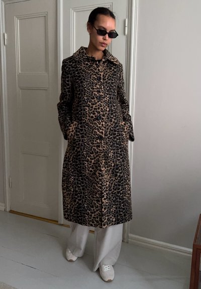 Mujer con un abrigo largo de estampado de leopardo, pantalones blancos de pierna ancha, zapatillas blancas y gafas de sol negras estrechas, de pie en el interior junto a unas puertas cerradas.