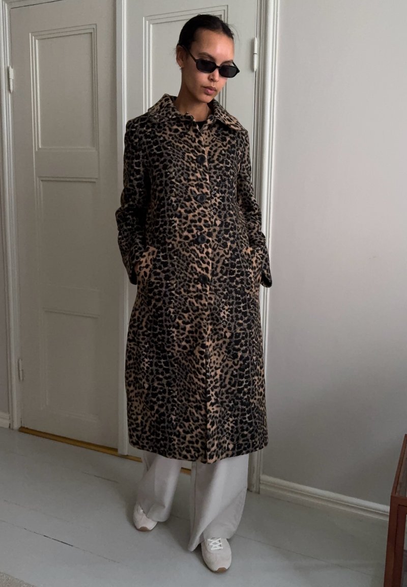 Sieviete, kas valkā garu leopardā rakstainu mēteli, baltas platajām kājām bikses, baltas sporta kurpes un melnus šaurus saulesbrilles, stāv telpās pie aizvērtām durvīm.