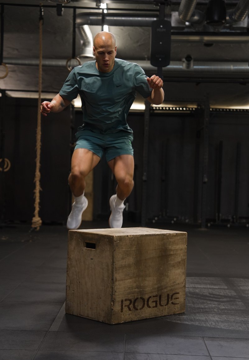 Homme en tenue de sport verte sautant sur une boîte plyo en bois dans une salle de sport avec des tuyaux industriels et du matériel en arrière-plan.